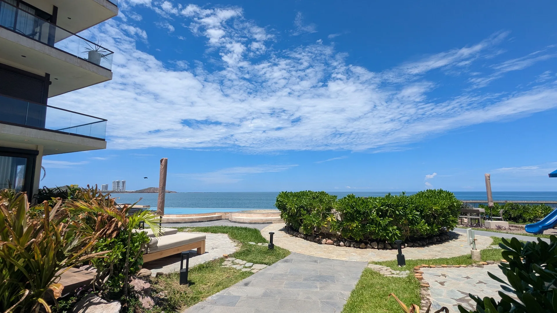 Andador de piedra entre jardines que lleva a una alberca frente al mar, con la costa de Mazatlán al fondo