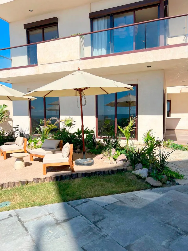 Rincón ajardinado Rincón exterior en planta baja de Aldea Ananta, con sombrilla, jardín y sillones de madera junto al edificio; un andador de piedra cruza el primer plano, con una segunda sombrilla idéntica más adelante.